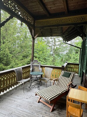 A covered outdoor area with wooden flooring, a lounge chair, a small table, and a chair.