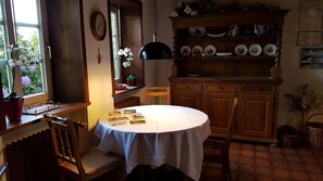 A dining area with a round table, chairs, a window with flowers, a clock, and a shelf with plates.