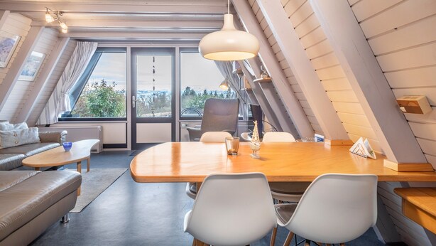 Et moderne loftrum med et træspisebord, hvide stole, en sofa og et stort vindue med udsigt over byen.