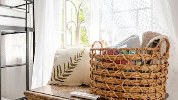 Un panier en osier est posé sur une table, à côté d’une télécommande, près d’une fenêtre ornée de rideaux légers.