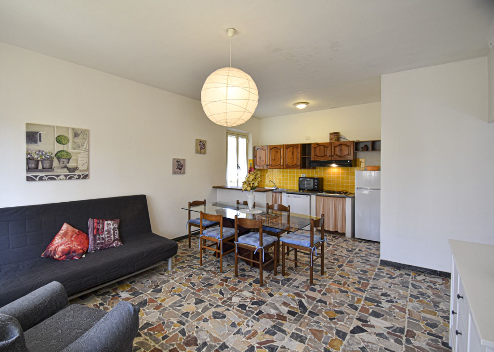 A living room with a sofa, a dining table with chairs, a kitchen with cabinets and appliances, and a mosaic floor.