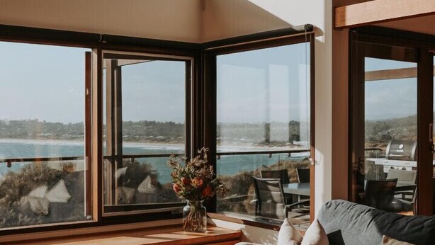 A modern living room with a large window offering a view of the sea and a patio area with outdoor furniture.