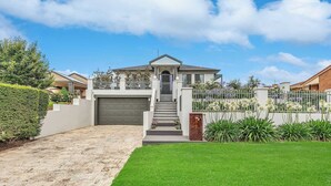 A modern two-story house with a front garage, a well-maintained lawn, and a decorative fence with flowering plants. - The Port Terrace (Echuca)