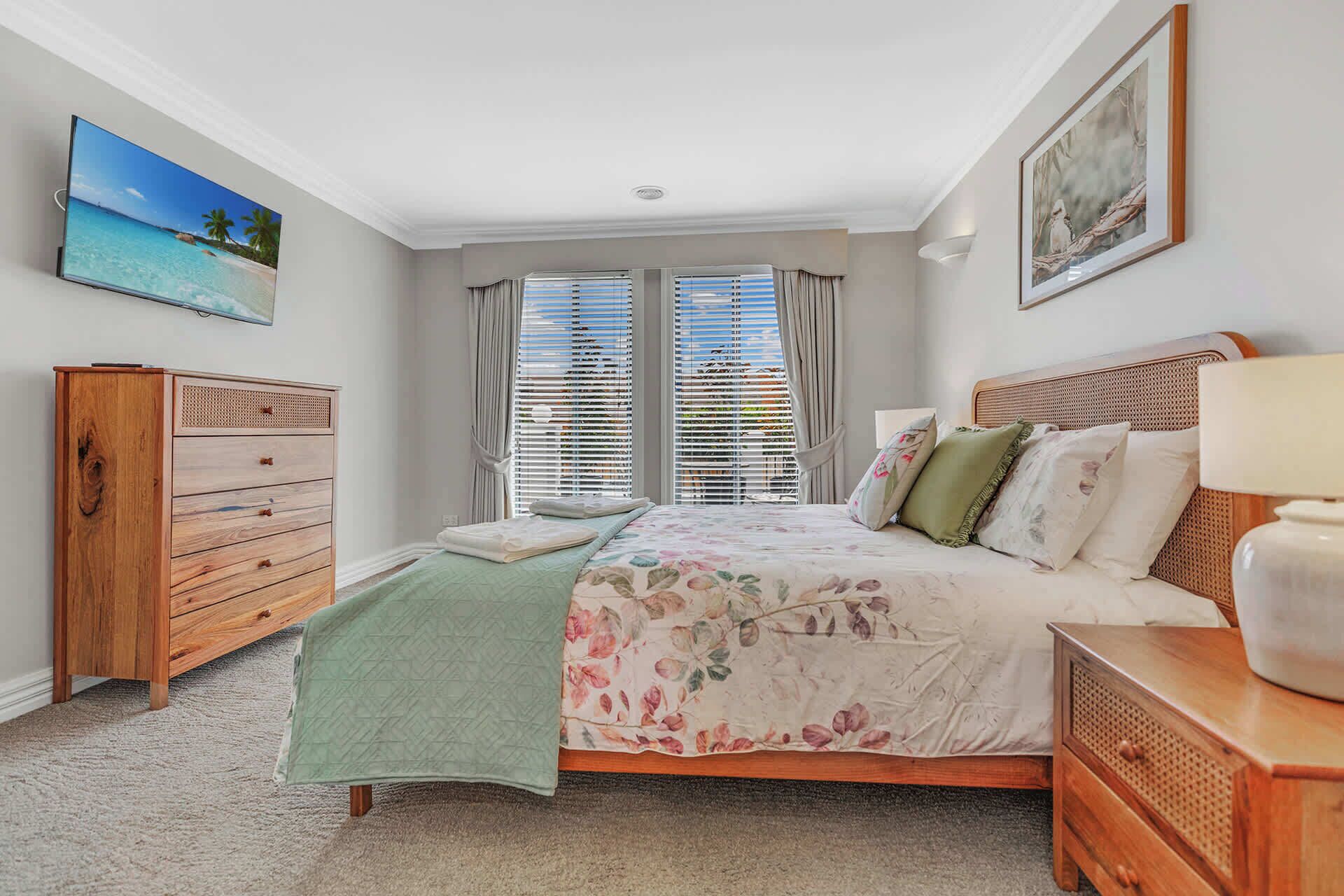 A bedroom with a bed, a wooden dresser, a nightstand, a painting, and a window with blinds.