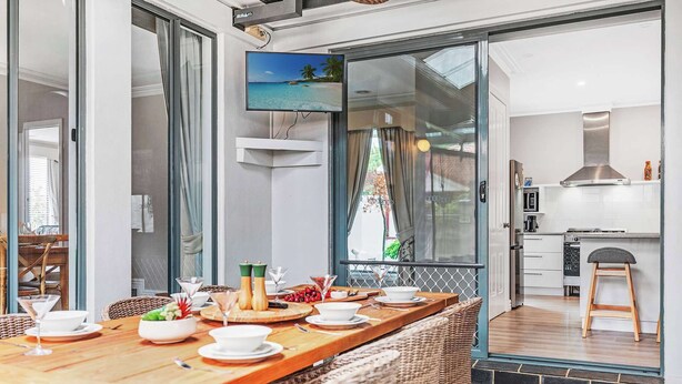 A dining area with a wooden table set for four, a television mounted above the table, and a kitchen visible through an open door. - The Port Terrace (Echuca)