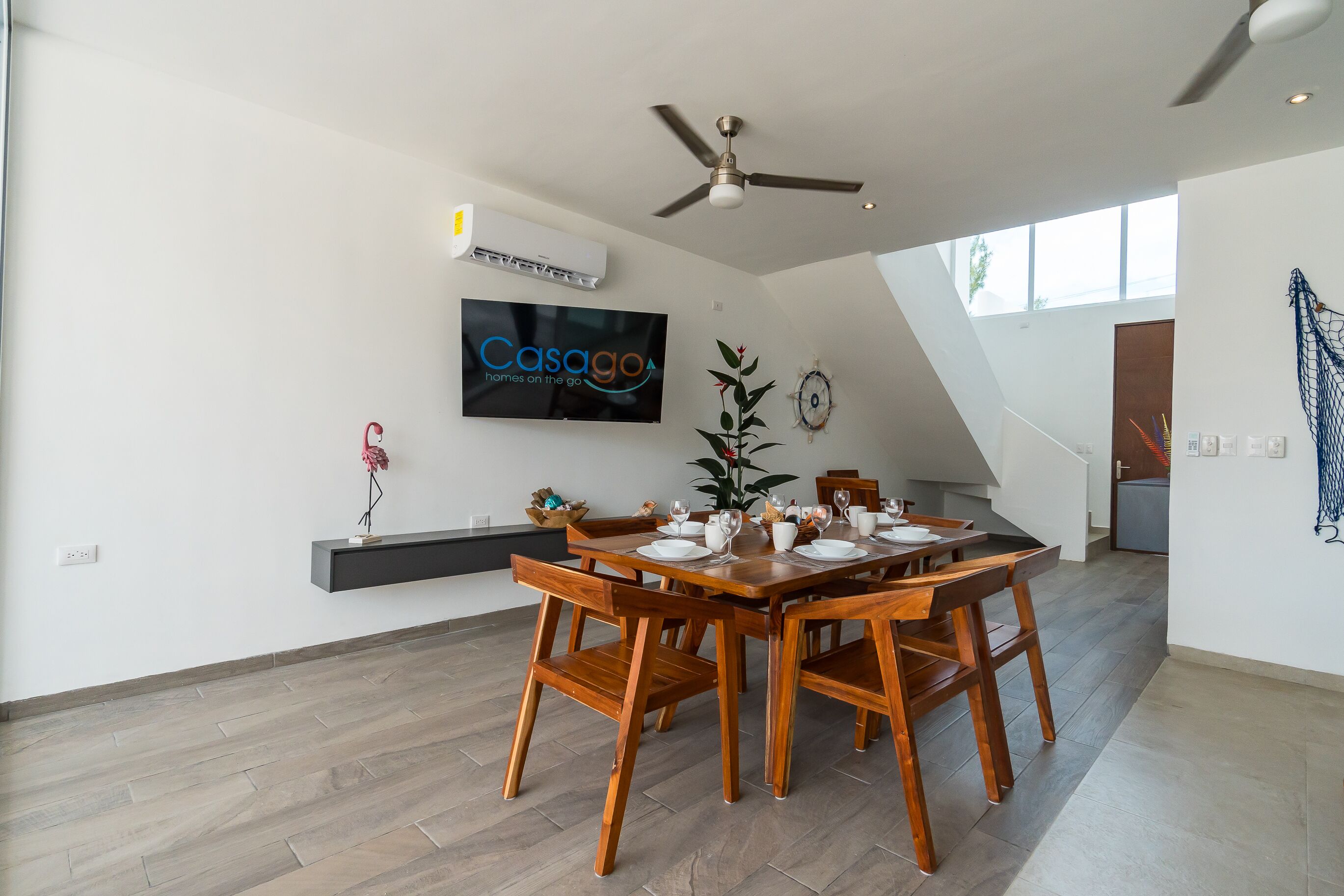 A modern dining area with a wooden table set for four, a ceiling fan, and a mounted TV displaying 'CasaGo'.