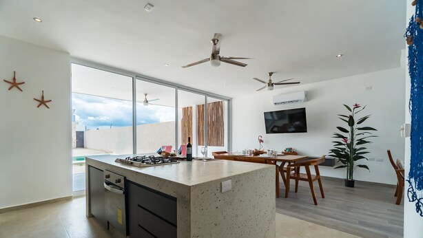Cocina moderna con una isla central, comedor con sillas de madera, ventiladores de techo y un amplio ventanal con vistas al cielo.