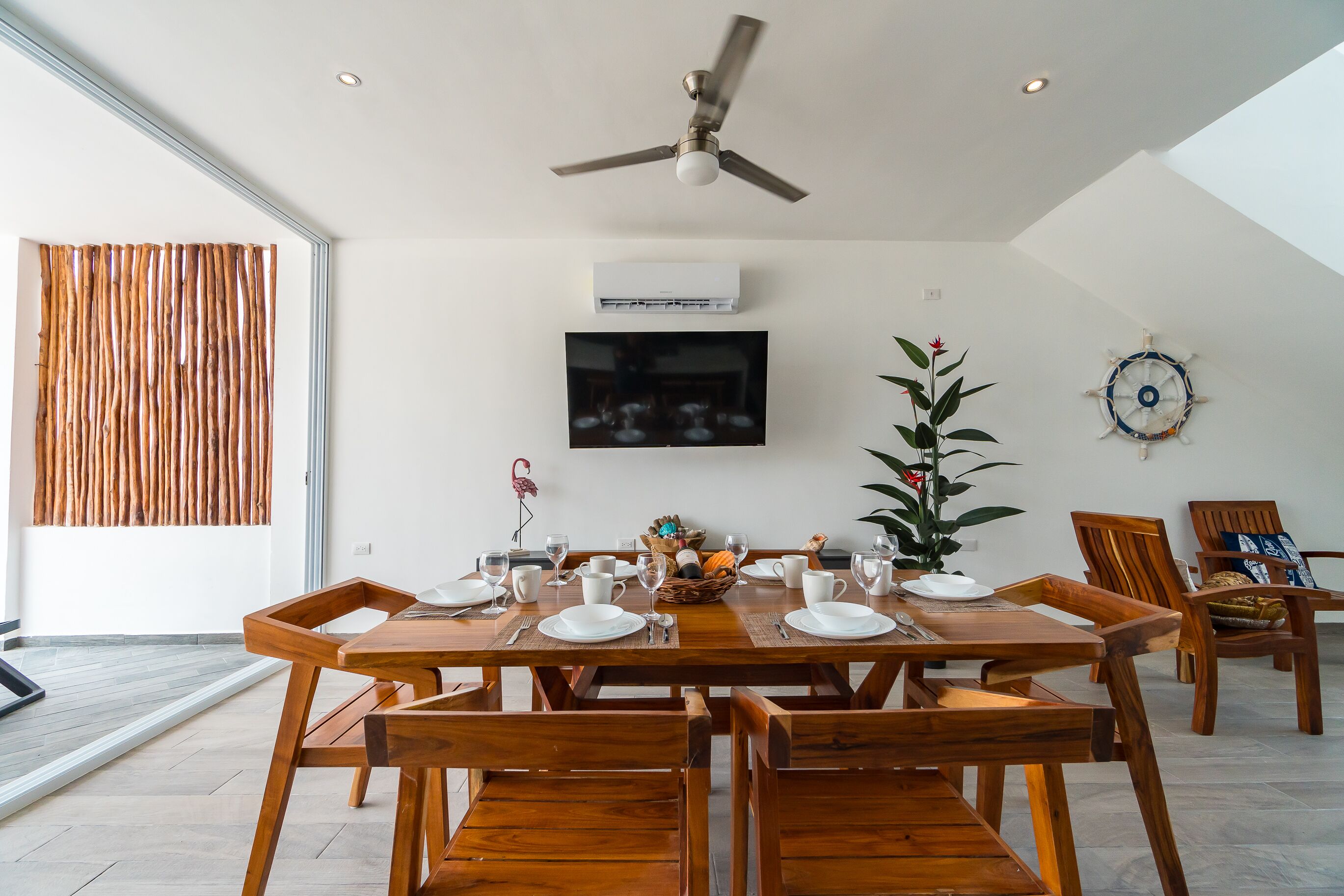 A dining area with a wooden table set for four, a ceiling fan, an air conditioning unit, a mounted television, and a decorative wall piece.