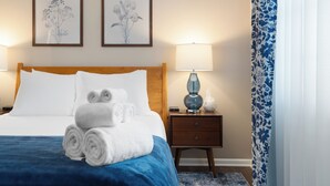 Een netjes opgemaakt bed met opgerolde witte handdoeken, een houten hoofdbord, twee ingelijste bloemrijke kunstwerken aan de muur, een bedlamp en een gordijn met dessin.