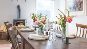 A dining room with a wooden table set for six, surrounded by wicker chairs. A fireplace is visible in the background, and there are floral centerpieces on the table. - Stone Barn (Skibbereen)