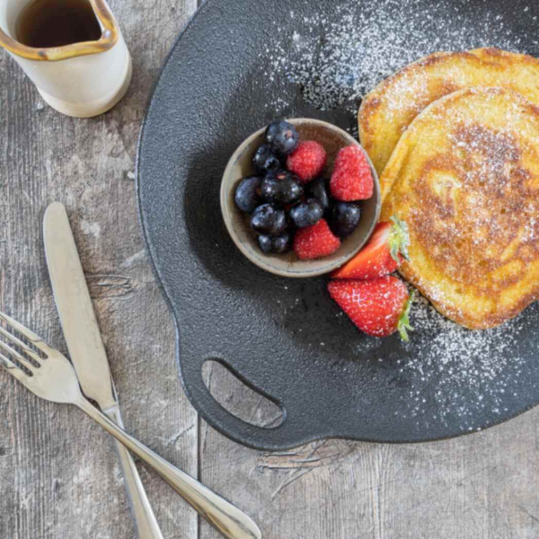 A plate with pancakes, blueberries, and strawberries, accompanied by a fork and knife on a wooden surface.
