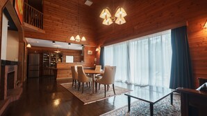 A spacious room with a wooden ceiling, a dining area with chairs, a kitchen in the background, and a large window with curtains.