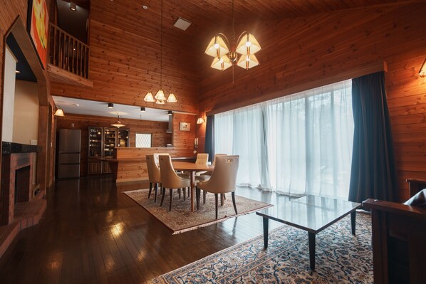 Una habitación amplia con techo de madera, un comedor con sillas, una cocina al fondo y un gran ventanal con cortinas.