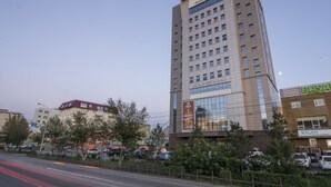 Un hotel de varios pisos con fachada de vidrio moderna, un letrero de restaurante visible y un estacionamiento con automóviles.