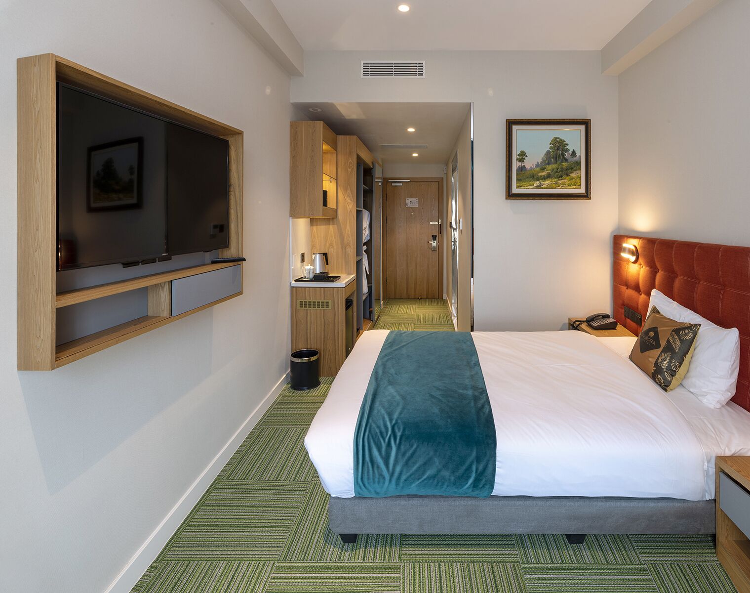 Habitación de hotel con cama, televisor montado en la pared, una kitchenette pequeña y un pasillo que conduce a otra habitación.