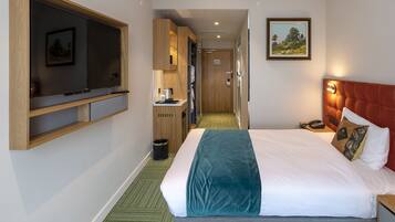 Habitación de hotel con cama, televisor montado en la pared, una kitchenette pequeña y un pasillo que conduce a otra habitación.