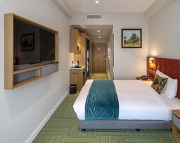 Habitación de hotel con cama, televisor montado en la pared, una kitchenette pequeña y un pasillo que conduce a otra habitación.