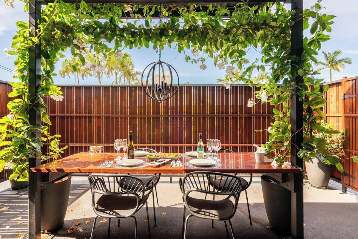 Une table en bois, dressée pour deux personnes, placée sous une pergola couverte de plantes grimpantes, et entourée d’une clôture en bois et de plantes en pot.
