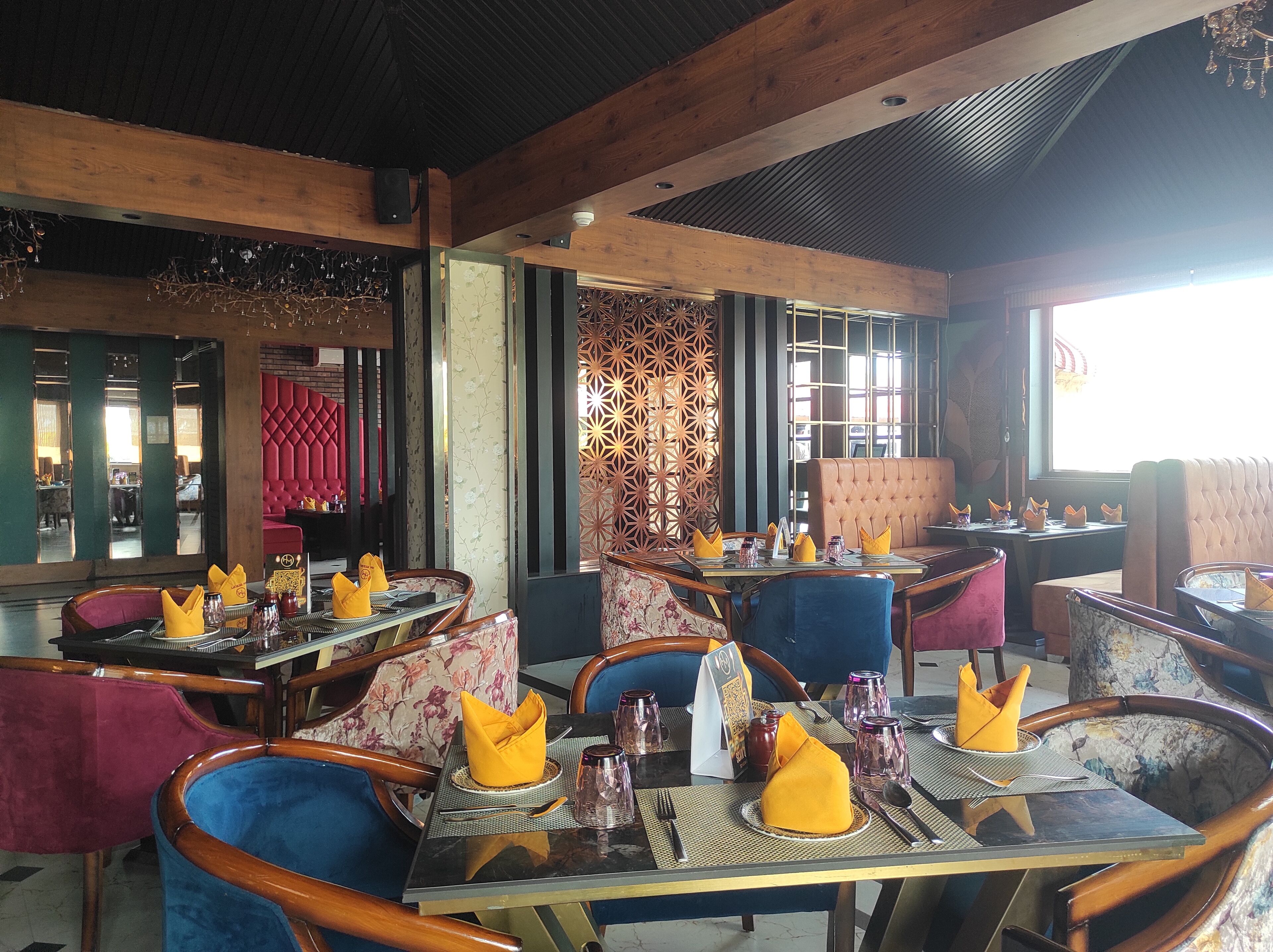 A restaurant interior with wooden beams, cushioned seating, and a patterned wall. Tables are set with yellow napkins and glassware.
