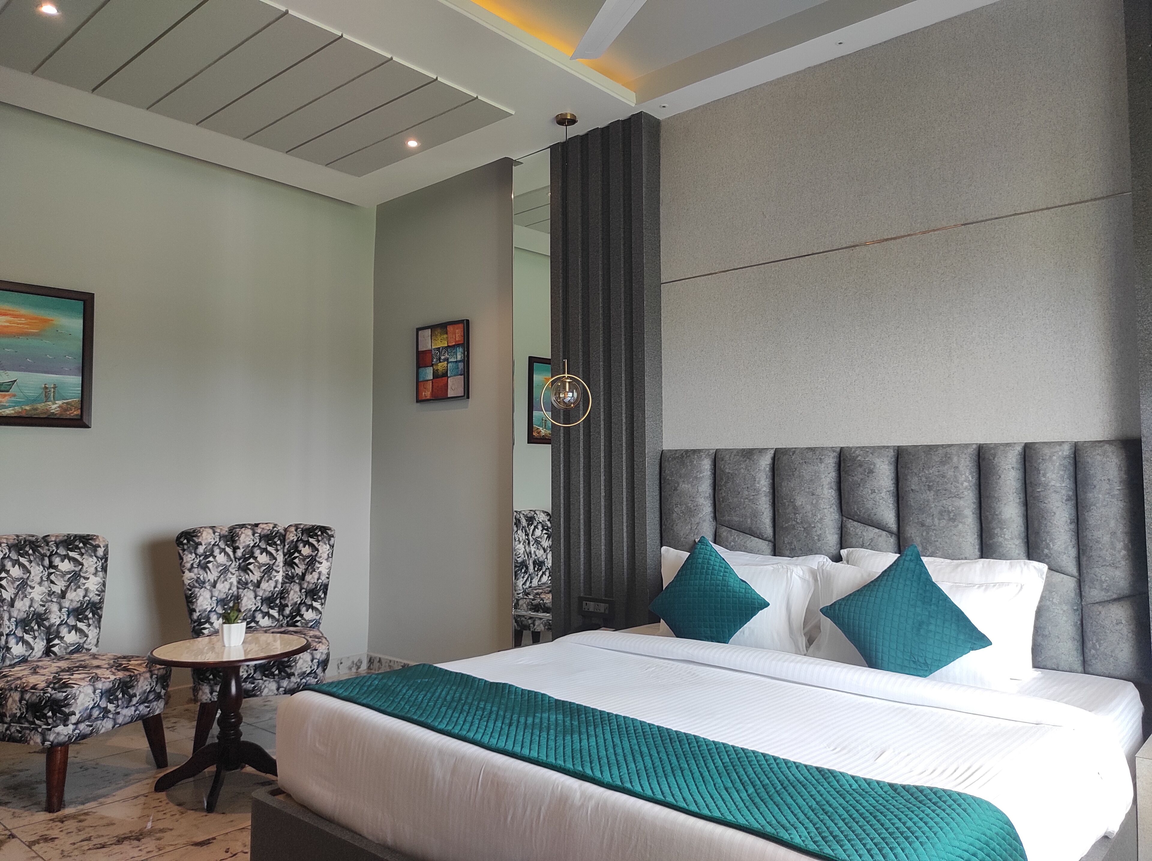 A modern hotel room with a large bed, two matching armchairs, a small round table, and a large window.