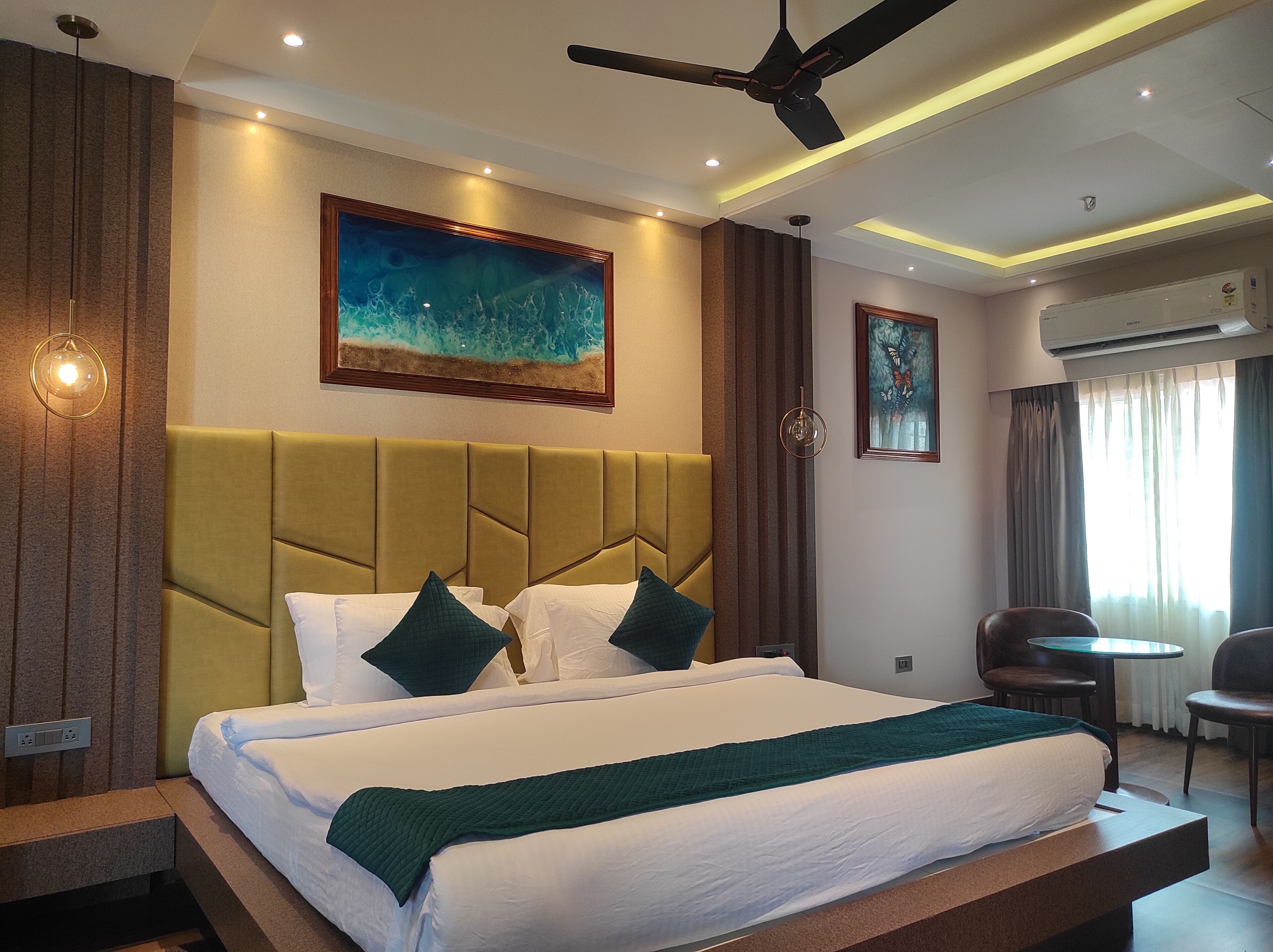 A hotel room with a large bed, a ceiling fan, a painting, an air conditioner, and a seating area with a table and chairs.