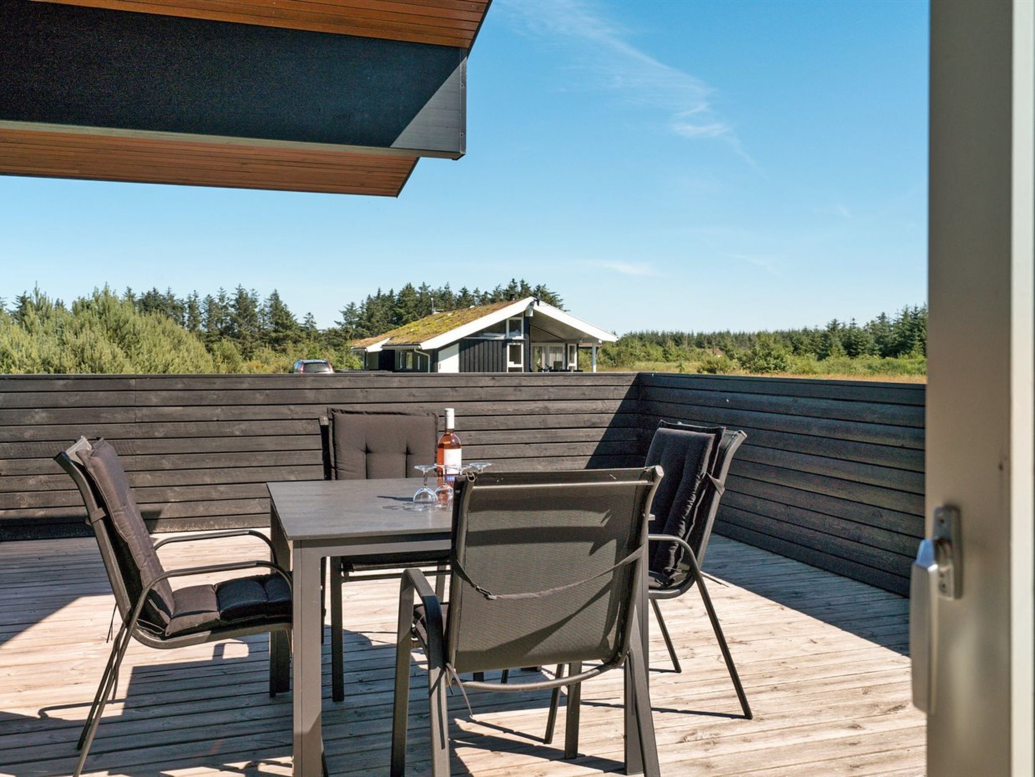 A wooden deck with a table and chairs, overlooking a house and forested area.