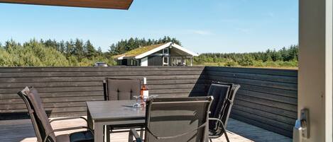 A wooden deck with a table and chairs, overlooking a house and forested area.