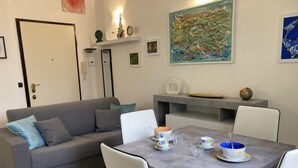Un salon moderne avec un canapé gris, une table à manger avec des chaises et une table basse. On trouve des objets décoratifs sur les étagères et un tableau au mur.