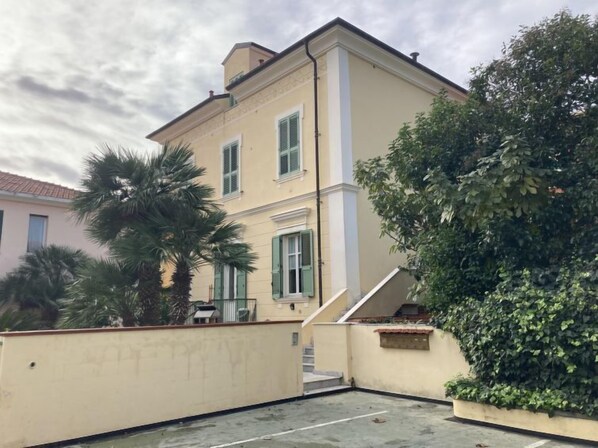 Um edifício de dois andares com fachada bege, persianas verdes e uma escadaria visível que leva ao andar superior.