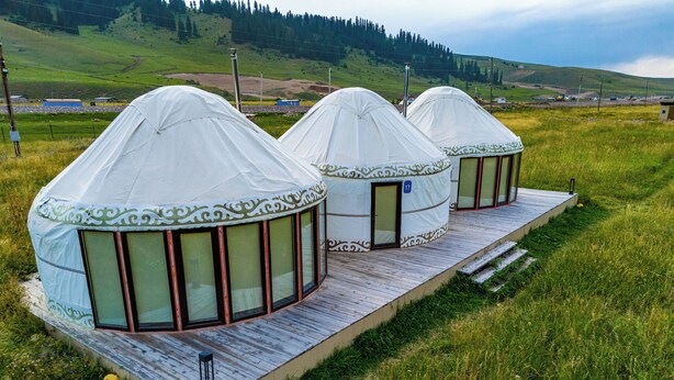 Three yurts with a wooden deck and large windows, set in a grassy field with a hilly background.
