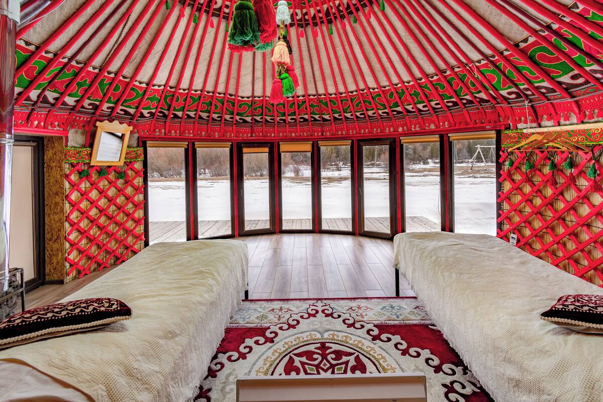 A traditional yurt with two beds, a red patterned ceiling, and a view of a snowy landscape.