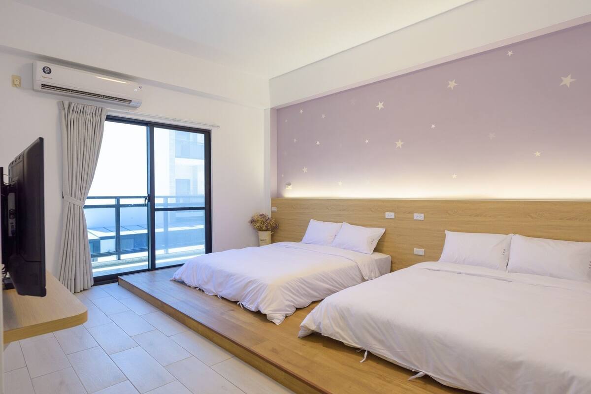 A hotel room with two beds, a TV, a large window with curtains, and a wall with star decorations.