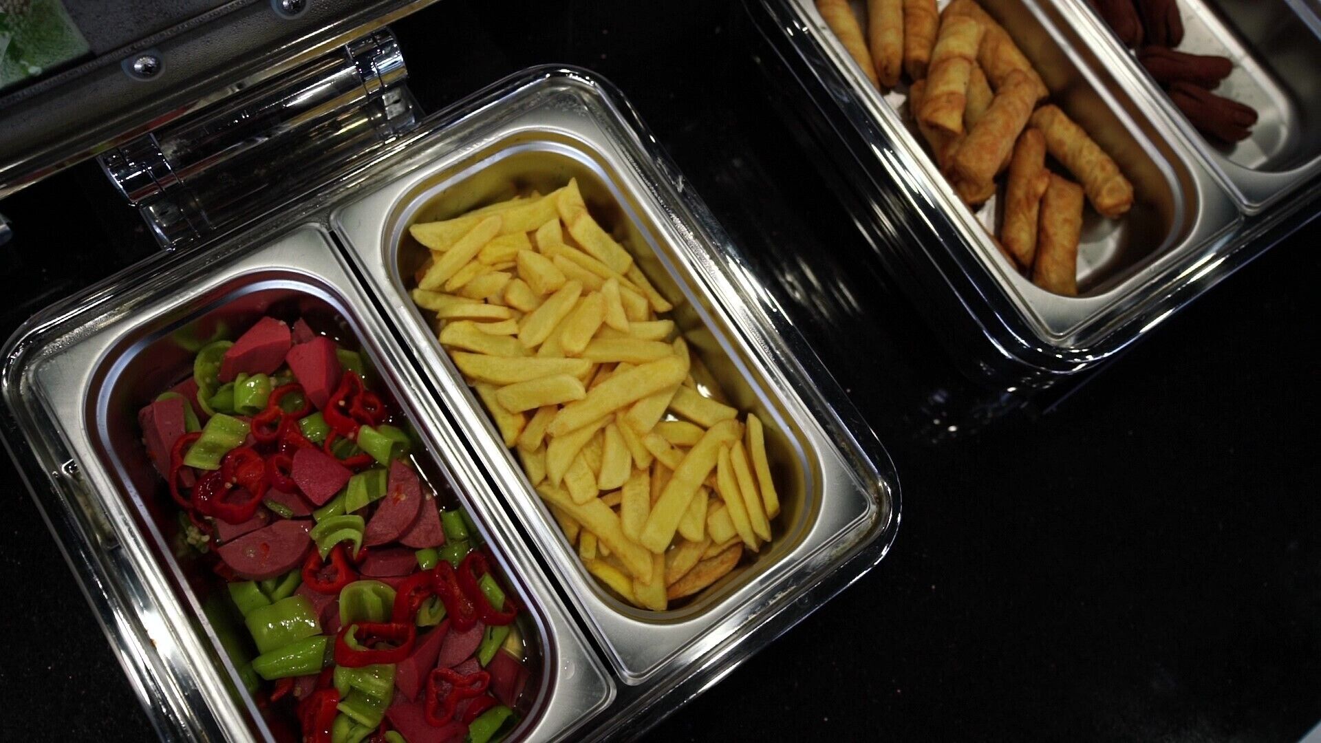 Un bufé con tres bandejas que ofrecen diferentes alimentos: una con pimientos rojos y verdes en rodajas, una con papas fritas y otra con rollitos fritos.