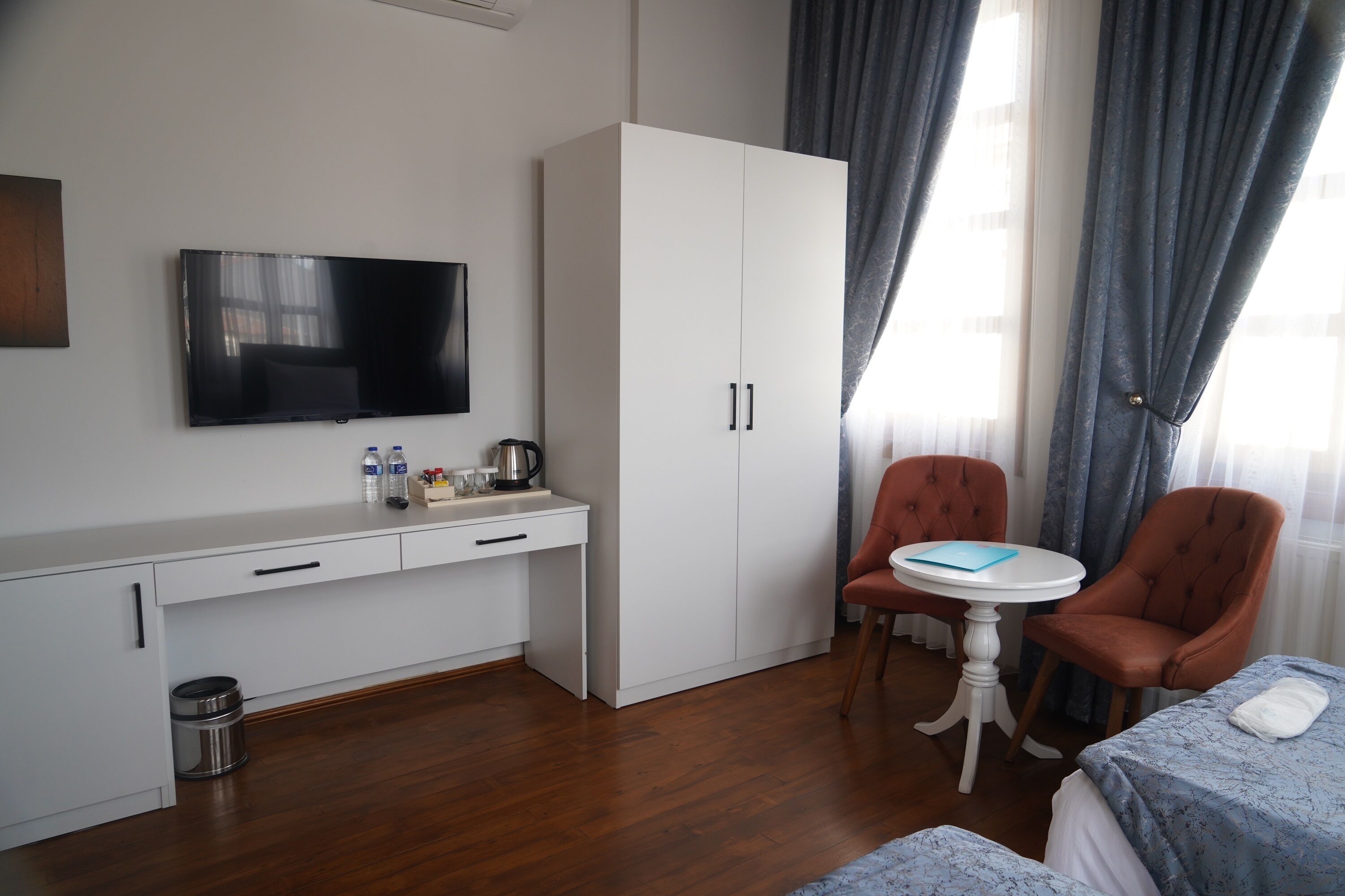 A hotel room with a flat-screen TV, a white wardrobe, a small table with a blue folder, two orange chairs, and a bed with a blue bedspread.