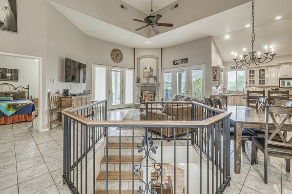 A spacious living area with a high ceiling, a fireplace, a dining area with a chandelier, and a staircase leading to an upper level.