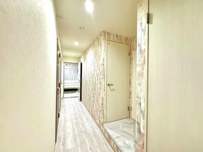 A narrow hallway with a door at the end, a small bench, and a bed visible through a window. - 450 (Osaka)
