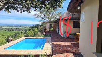A pool area with a view of the countryside, a patio with a table and chairs, and a building with a thatched roof.