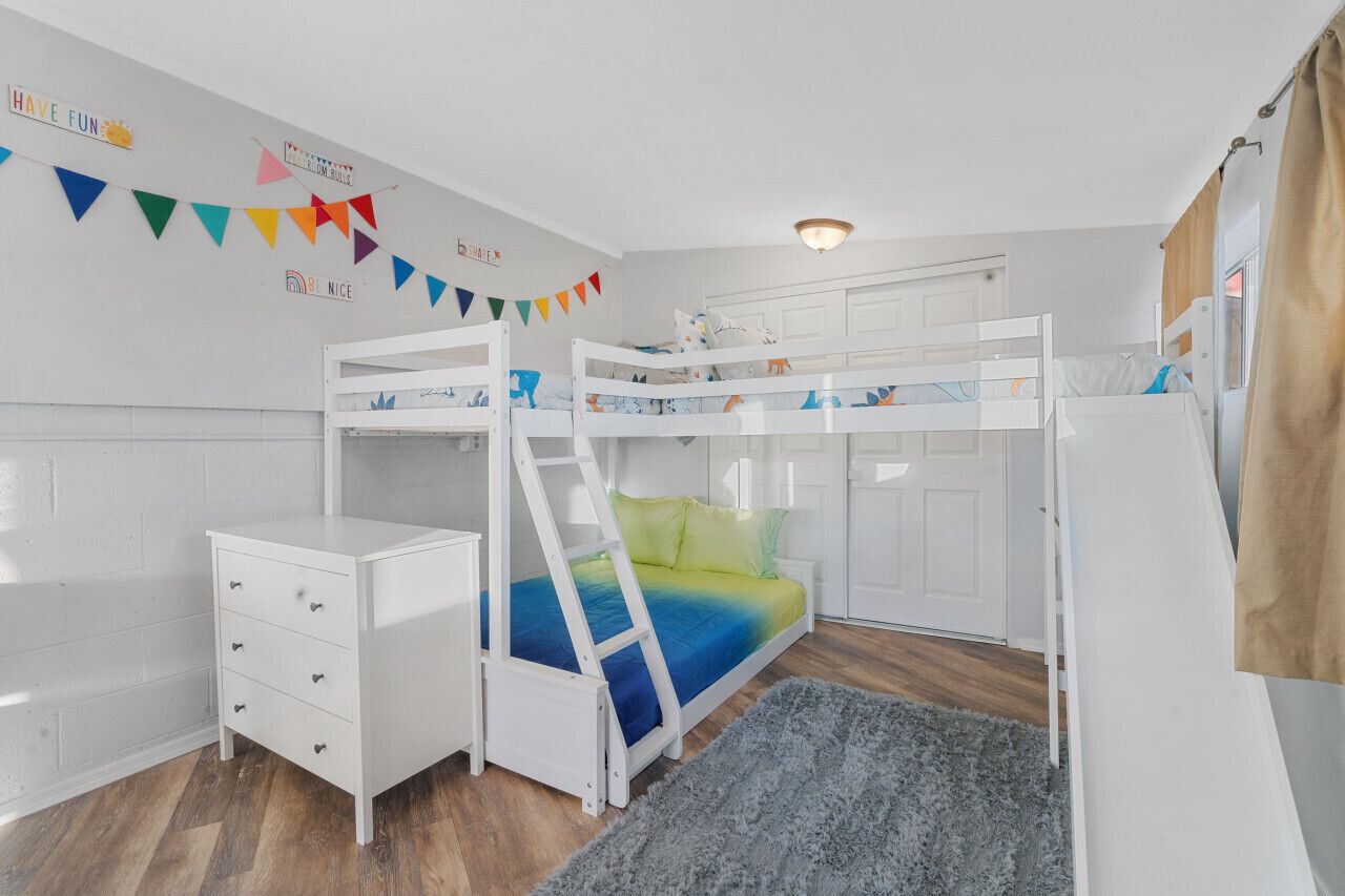 Quarto de criança com beliches, cômoda branca, tapete cinza e decorações coloridas nas paredes.