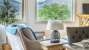 A cozy living room with a wooden coffee table, a chair with a blue throw, a lamp, and a view of mountains through the windows. - "Rundle Vista" at Canmore Crossing (Canmore)