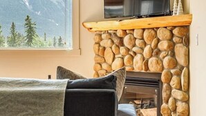 A cozy living room with a stone fireplace, a television mounted above it, and a view of mountains through a large window. - "Rundle Vista" at Canmore Crossing (Canmore)