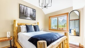 A bedroom with a wooden bed frame, a large window with a mountain view, a painting on the wall, and a chandelier. - "Rundle Vista" at Canmore Crossing (Canmore)