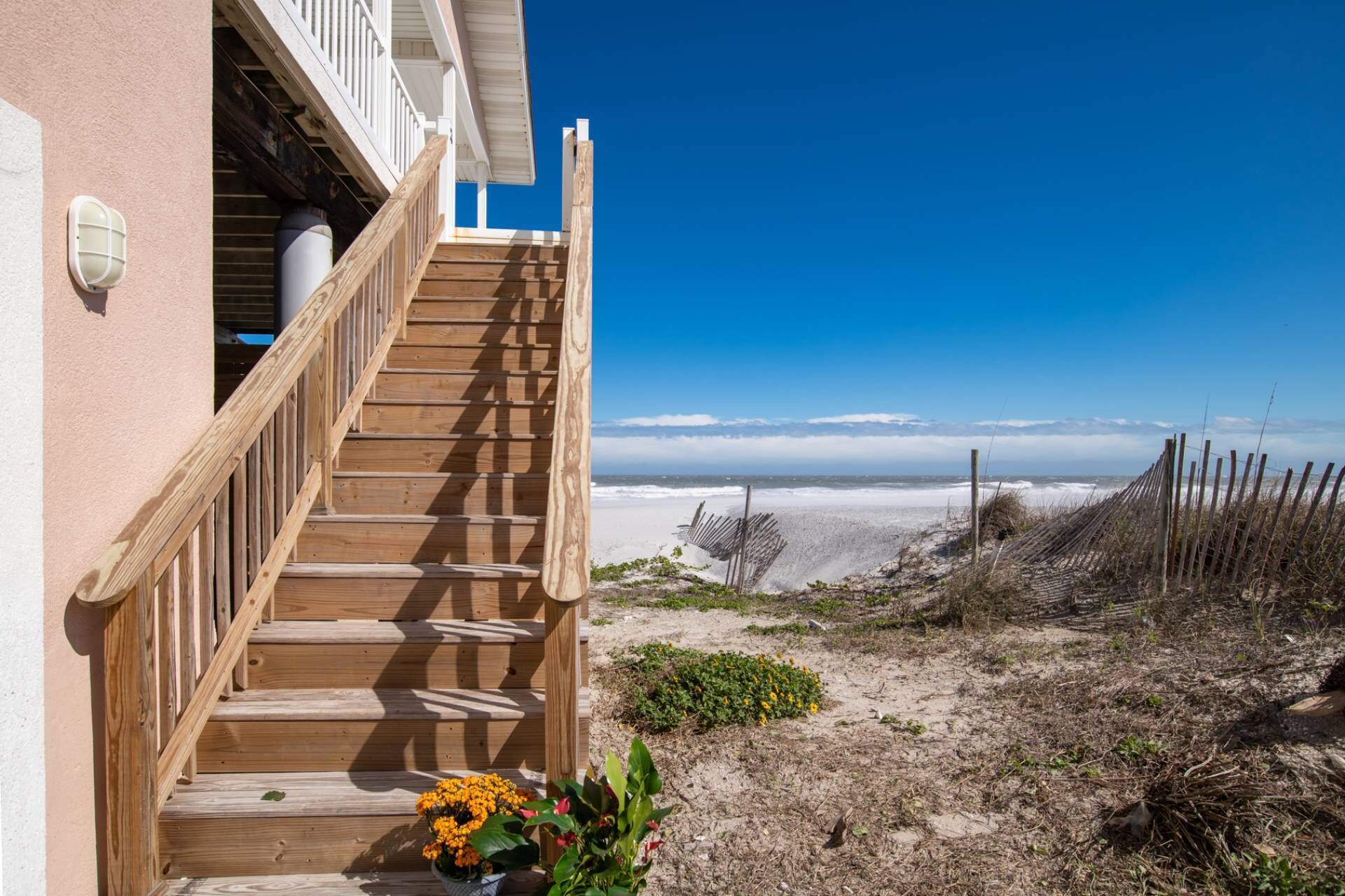 A wooden staircase leading to a balcony overlooking a beach with a fence and some vegetation.