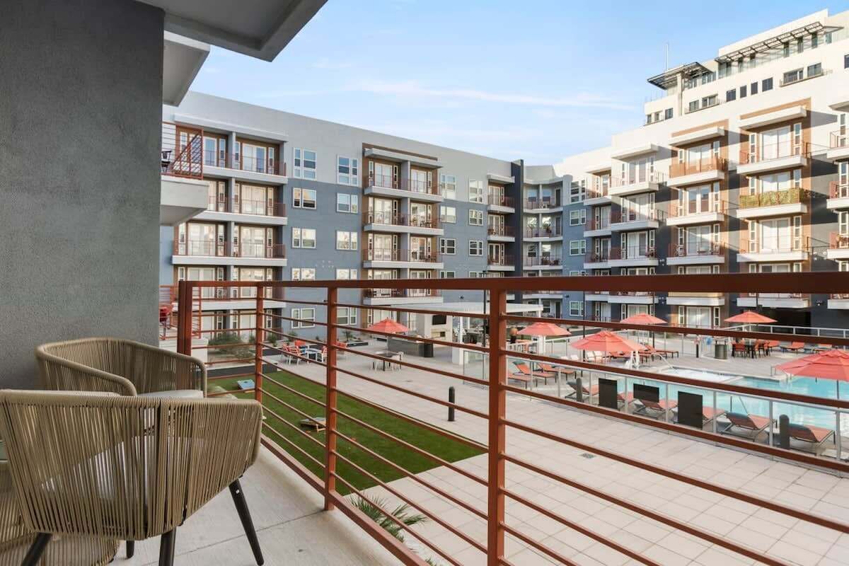Un balcone con sedie in vimini, una zona piscina con ombrelloni e vista su edifici a più piani.