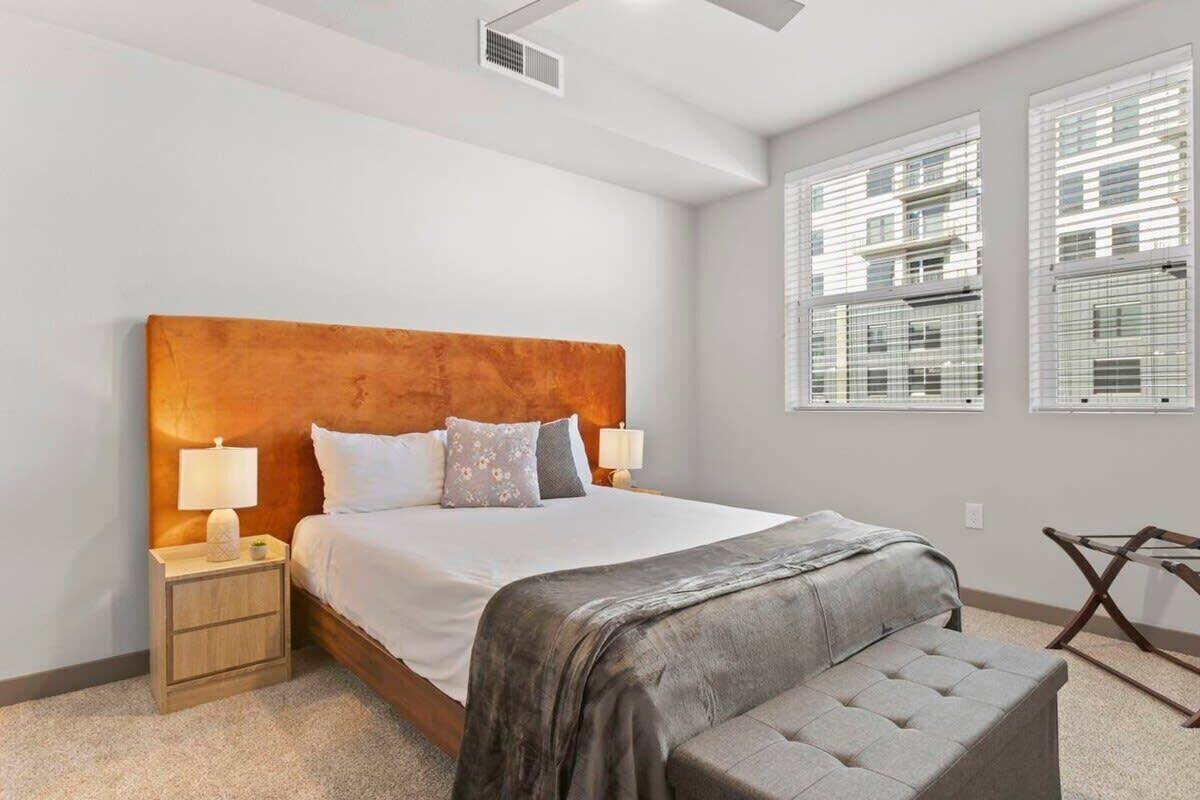 A modern hotel room with a large bed, two bedside lamps, a grey bench, and a view of buildings through the windows.