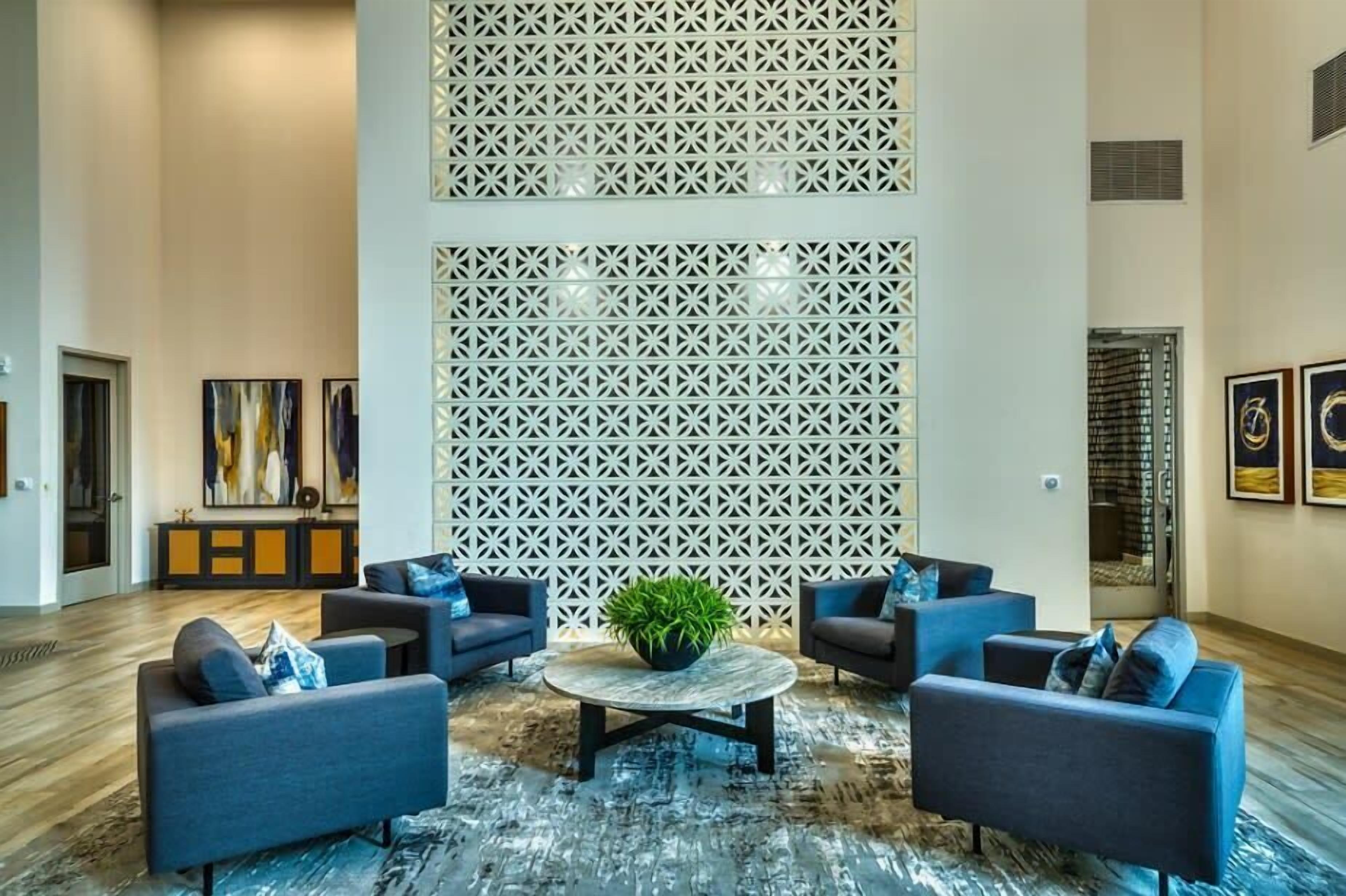 A spacious lobby with a modern design, featuring a central round table with a potted plant, surrounded by sofas. A decorative white lattice wall and artwork are visible.