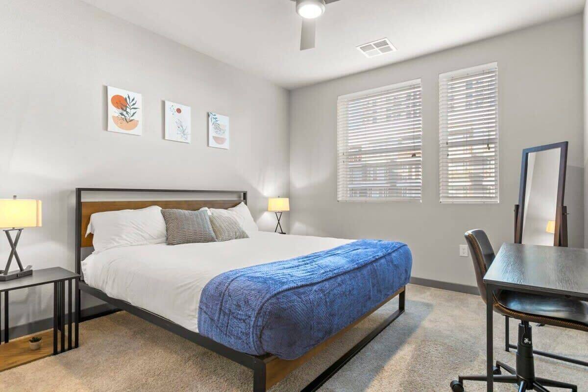 A bedroom with a bed, bedside tables, a desk, and a chair. There are three wall-mounted pictures and two windows with blinds.