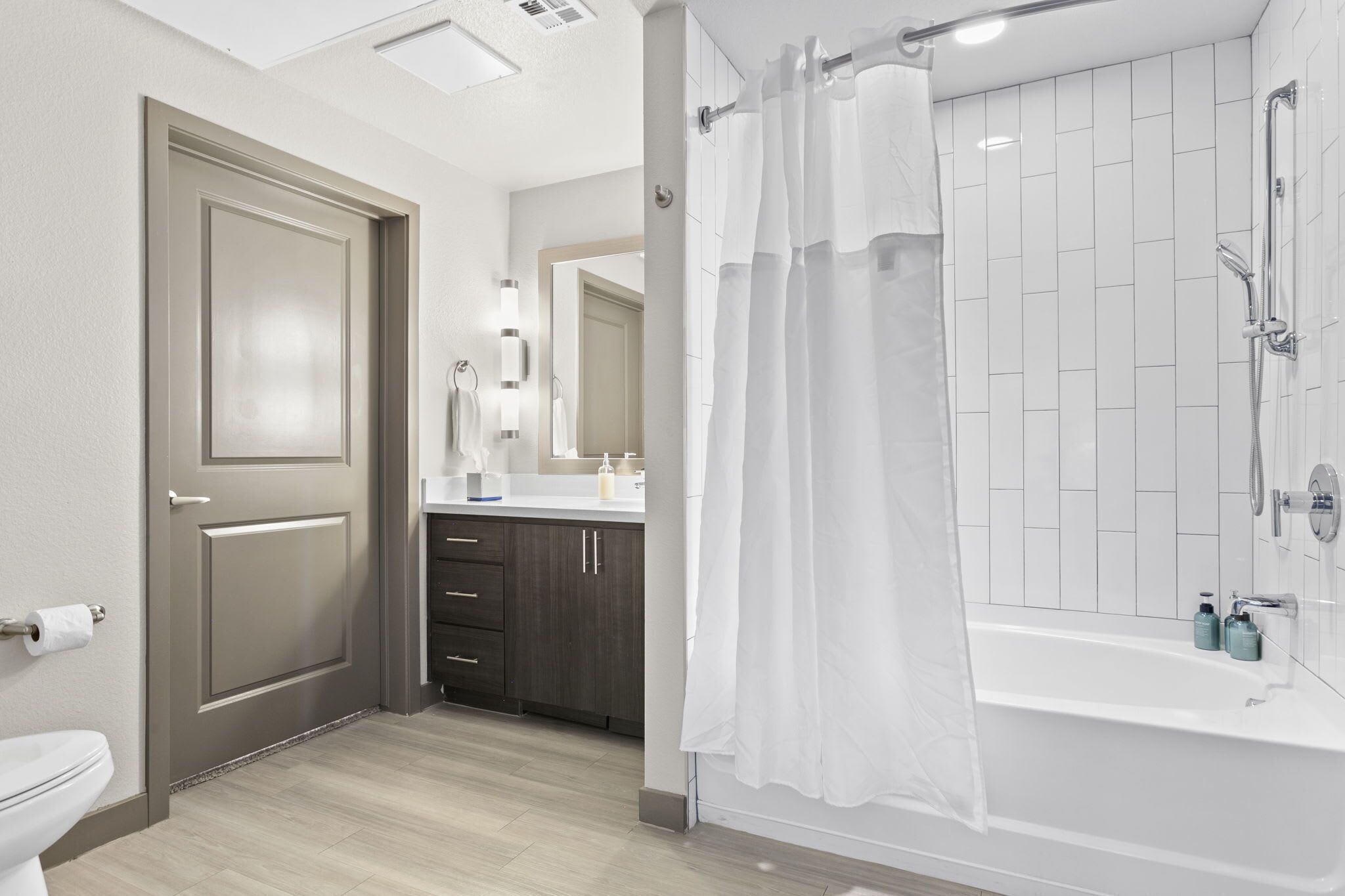 A modern bathroom with a toilet, dark wooden cabinets, a white bathtub with a shower curtain, and a mirror above the sink.