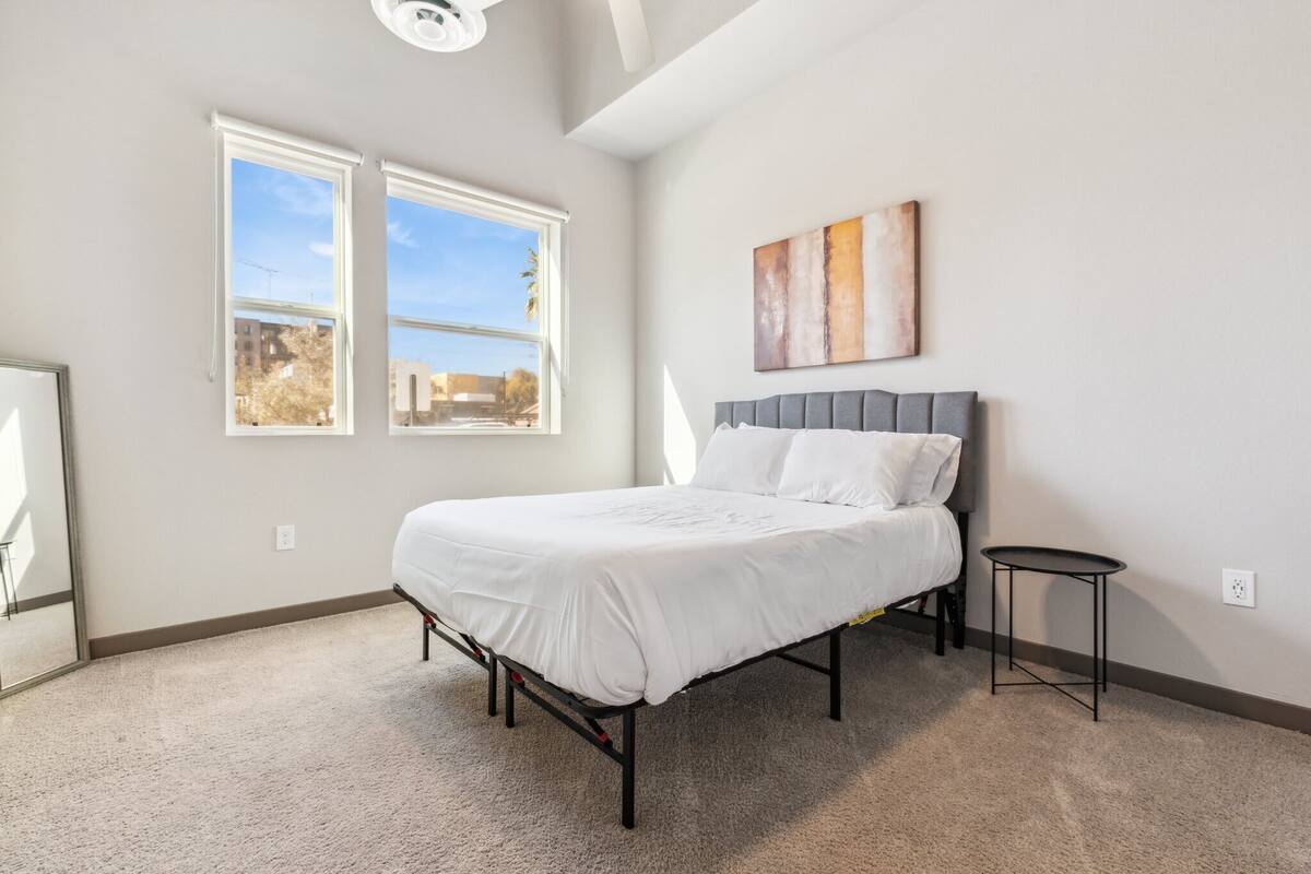 A bedroom with a bed, two windows, a small table, and a wall-mounted artwork.