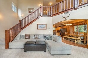 A spacious living room with a large sectional sofa, a wooden staircase, and a kitchen area visible through an open archway. - PET-FRIENDLY FOREST RETREAT CABIN | HOT TUB, FIRE-PIT, & A/C (Duck Creek Village)