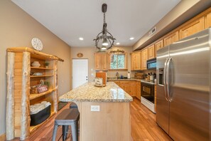 A kitchen with wooden cabinets, a granite countertop, stainless steel appliances, and a hanging light fixture. - PET-FRIENDLY FOREST RETREAT CABIN | HOT TUB, FIRE-PIT, & A/C (Duck Creek Village)